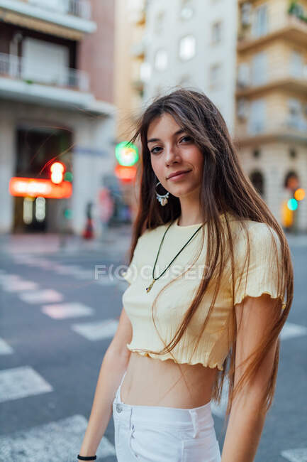 Vista laterale della giovane donna gentile che guarda la fotocamera contro la strada urbana alla luce del sole morbida — Foto stock