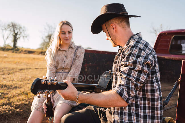 Novio en sombrero de vaquero tocando la guitarra acústica mientras está sentado con su novia en el maletero de coche rojo retro pickup estacionado en la carretera de arena en el campo - foto de stock