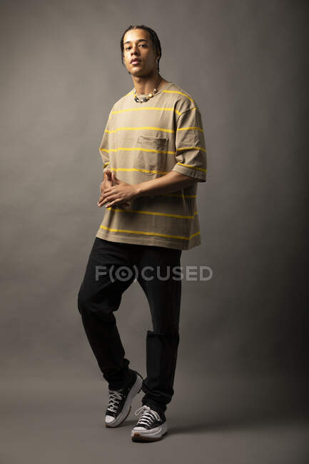 Joven modelo masculino afroamericano con cabello trenzado vestido con camisa y collar de rayas de gran tamaño mirando a la cámara sobre fondo gris - foto de stock