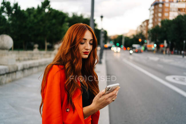 Smiling female with red hair and in orange suit text messaging on mobile phone while walking in city street — Stock Photo