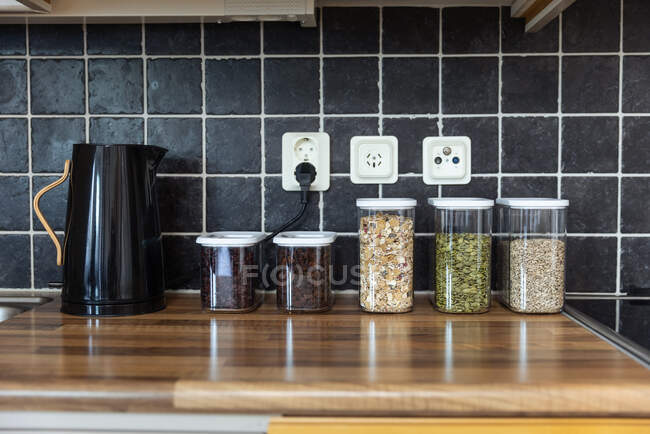Recipientes de plástico com muesli e sementes de girassol e abóbora colocados perto de grãos de café e chaleira elétrica no balcão na cozinha — Fotografia de Stock
