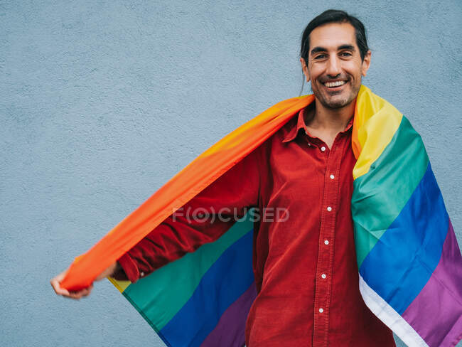 Gay ethnic male wrapped in rainbow LGBT flag looking at camera against gray wall in city — Stock Photo