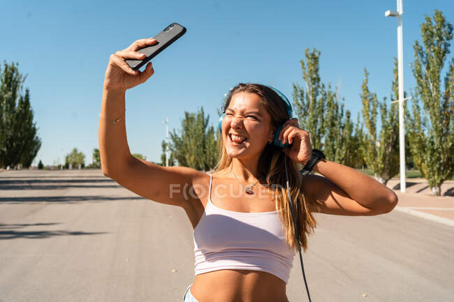 Patinadora femenina positiva en patines y auriculares tomando fotos en el teléfono móvil parpadeando y sacando la lengua en un día soleado en verano en la ciudad - foto de stock