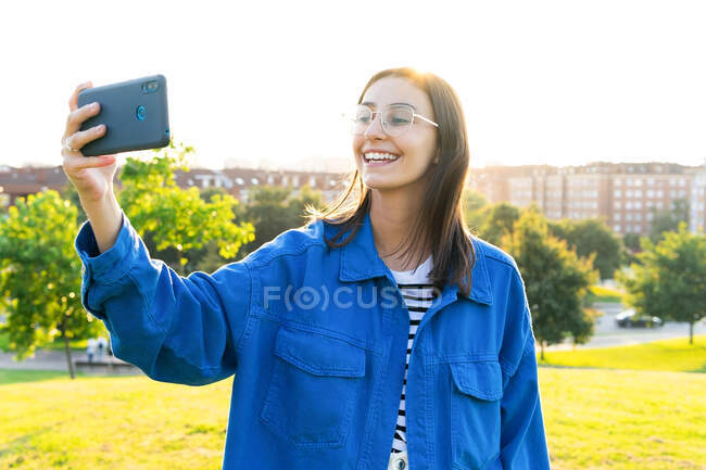 Stylish female standing on green grassy hill and taking picture of cityscape on smartphone on sunny day — Stock Photo