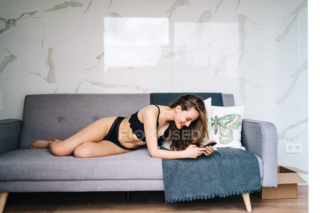 Deliziosa donna in biancheria intima nera sdraiata sul divano e messaggistica sui social media tramite telefono cellulare in accogliente salotto a casa — Foto stock