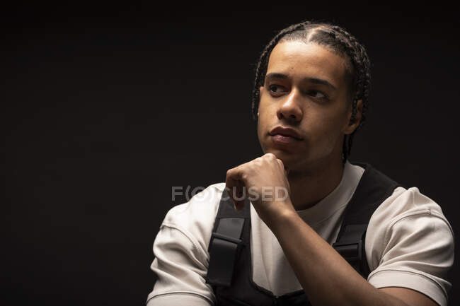 Красивый молодой этнический мужчина с афрокосичками, одетый в черно-белую одежду, смотрит в сторону, сидя в темной студии — стоковое фото