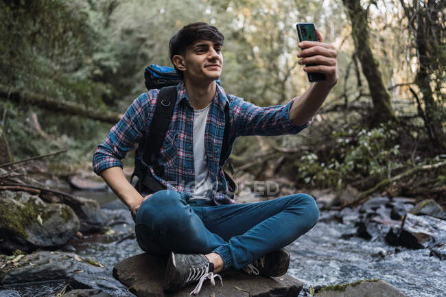 Senderista masculino sonriente tomando uno mismo tiro en el teléfono inteligente mientras está sentado en el fondo del lago en los bosques durante el trekking - foto de stock