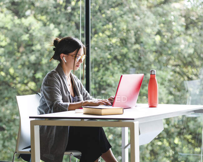 Seitenansicht einer Studentin, die am Netbook tippt, während sie mit Buch am Tisch sitzt und Hausaufgaben in der Universitätsbibliothek macht — Stockfoto