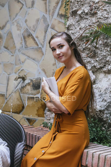 Seitenansicht der charmanten jungen Studentin, die mit Buch vor einer Steinmauer auf der Straße steht und in die Kamera blickt — Stockfoto