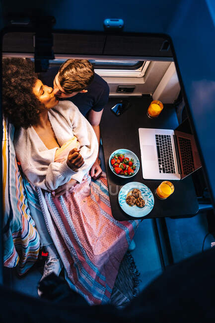 Von oben ein glückliches multiethnisches Paar, das sich beim Essen in der Nähe des Laptops sanft küsst, während es sich im Urlaub im Wohnmobil ausruht — Stockfoto