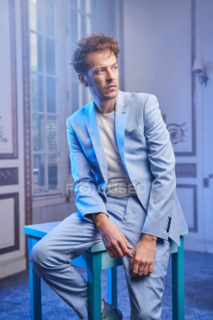 Pensativo hombre elegante en traje de mesa de estar en la habitación elegante y mirando hacia otro lado - foto de stock