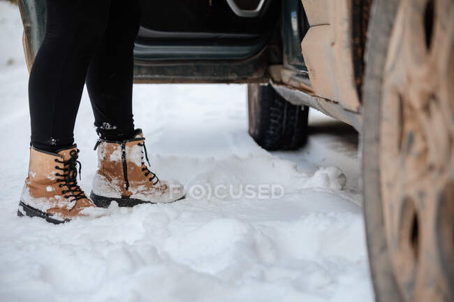 Crop anonymous female in warm clothes getting out of car parked on snowy road in winter woods — Stock Photo