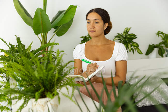Mujer étnica pensativa con pluma y agenda entre plantas verdes en macetas en el jardín de la casa - foto de stock