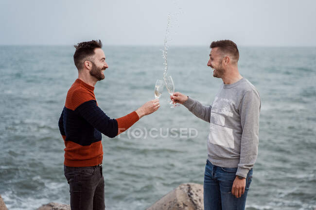 Vue latérale des hommes homosexuels gais cliquetis verres avec éclaboussure champagne tout en regardant les uns les autres contre l'océan — Photo de stock