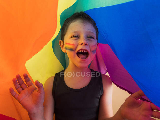 Criança positiva em camiseta com maquiagem nas bochechas e boca aberta gritando enquanto olha para longe sob bandeira LGBTQ — Fotografia de Stock