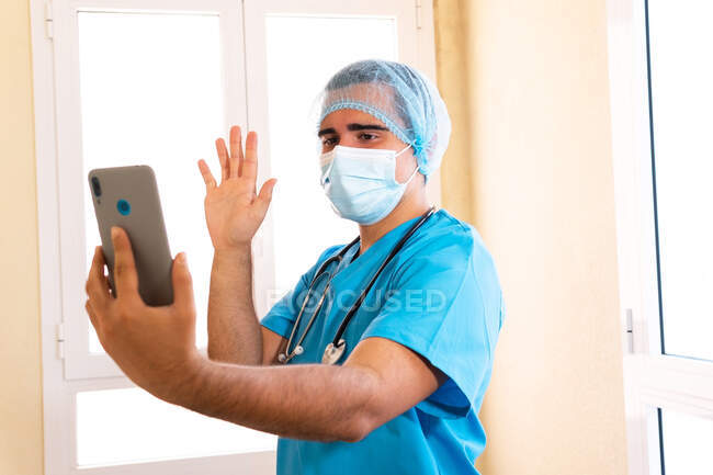 Medico maschile in maschera e uniforme mano ondulata e parlare in video chat tramite telefono cellulare in corridoio in clinica — Foto stock