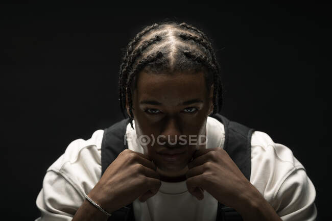 Seriöses junges afroamerikanisches Model mit geflochtener Frisur trägt stylisches Outfit und blickt vor schwarzem Hintergrund in die Kamera — Stockfoto