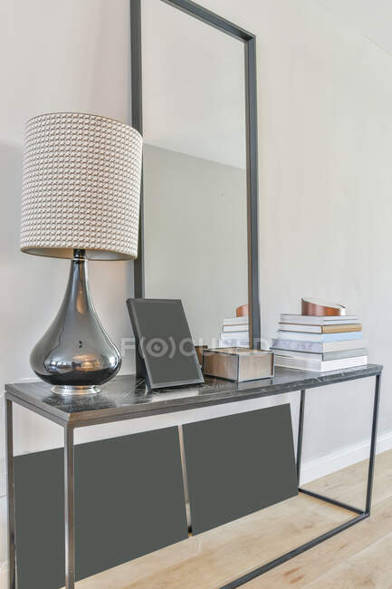 Современный дизайн интерьера дома со стильной лампой и рамкой, размещенной на полке из мрамора с книгами и зеркалом в современном светлом помещении — стоковое фото
