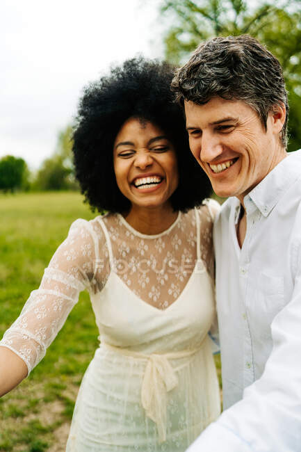 Cheerful multiracial couple stroking gray horse pasturing on meadow in countryside in summer — Stock Photo