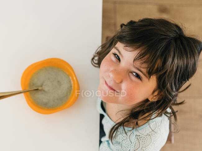 De cima de criança alegre sentada à mesa com tigela de sopa de creme e olhando para a câmera durante o almoço na cozinha em casa — Fotografia de Stock