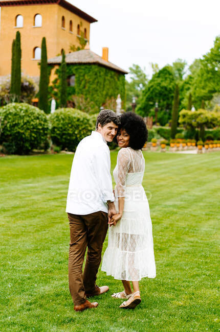 Fröhliches multiethnisches Paar hält Händchen und läuft im Garten gegen Gebäude, während er in die Kamera schaut — Stockfoto