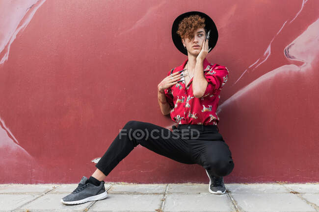Homem homossexual na moda com unhas compridas em camisa ornamental olhando para longe enquanto se agacha no pavimento contra a parede — Fotografia de Stock