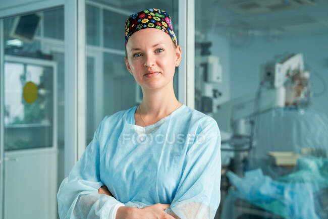 Médico adulto seguro de sí mismo con brazos cruzados en gorra médica ornamental mirando a la cámara contra la pared de vidrio en el hospital - foto de stock