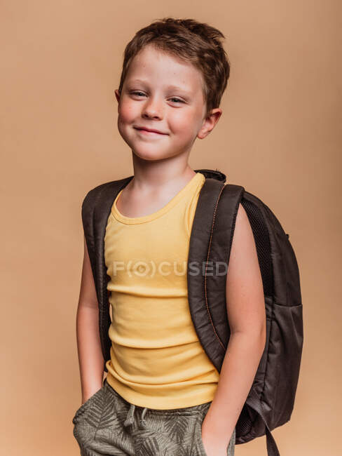 Positiv cooler Preteen-Schüler mit Rucksack blickt in die Kamera auf braunem Hintergrund im Studio — Stockfoto