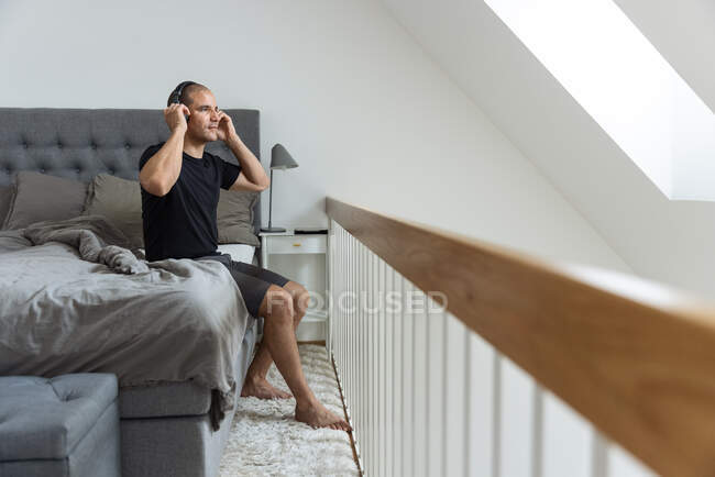 Вид збоку спокійного чоловіка одягає навушники і слухає музику, сидячи в ліжку після пробудження в спальні і насолоджуючись ранком — стокове фото