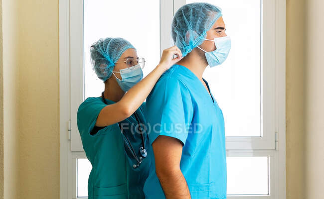 Side view of female medic helping male coworker putting on disposable medical cap while working in hospital — Stock Photo