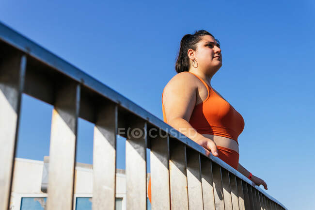 Vista lateral de atleta femenina contemplativa étnica con cuerpo curvo mirando hacia otro lado detrás de la valla bajo el cielo azul - foto de stock