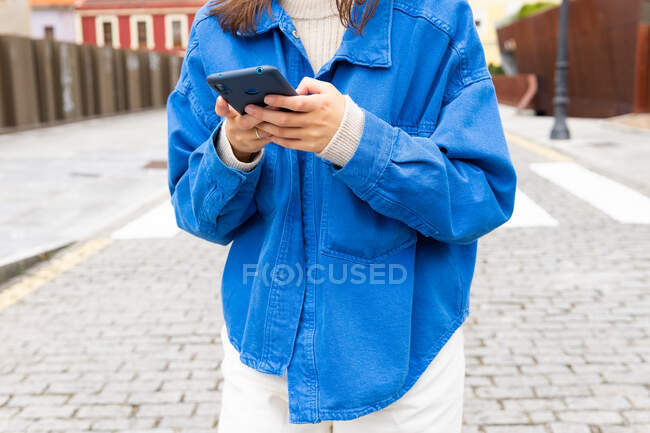 Анонімна жінка зі спантеличеним волоссям, що йде по вулиці міста в вітряний день і використовує смартфон — стокове фото
