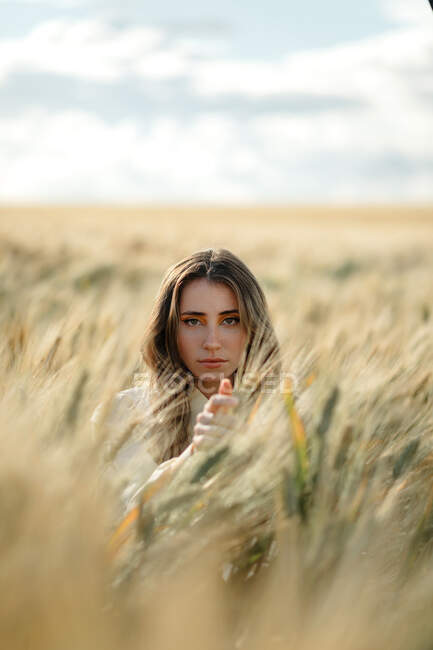 Mujer joven con el pelo ondulado mirando a la cámara en el campo bajo el cielo nublado sobre fondo borroso - foto de stock