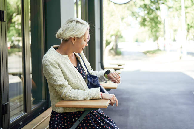 Vista lateral da mulher na moda sentada à mesa no café da rua e verificando o tempo no relógio de pulso enquanto espera pela nomeação — Fotografia de Stock