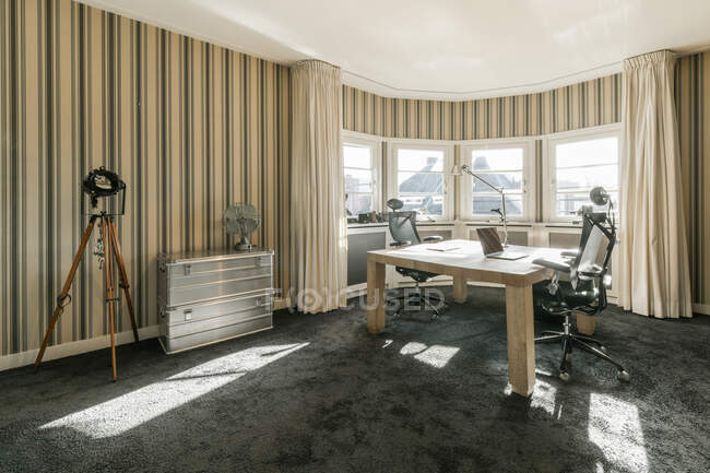 Mesa de madera con portátil y cómodos sillones colocados cerca de la ventana panorámica en una amplia habitación con papel pintado a rayas en un apartamento contemporáneo - foto de stock
