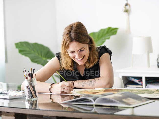 Umtriebige Designerin malt mit Pinsel auf Papier, während sie im Kreativbüro am Tisch sitzt und an einem Projekt arbeitet — Stockfoto