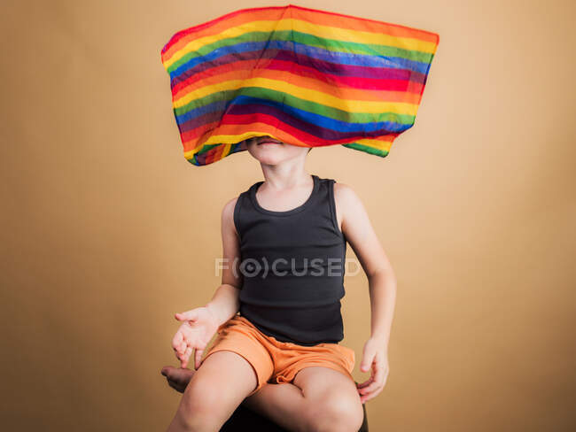 Anonymes barfüßiges Kind sitzt mit überkreuzten Beinen und Regenbogenfahne auf dem Kopf auf beigem Hintergrund — Stockfoto