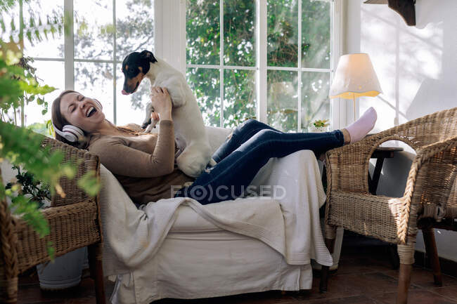 Vista lateral da fêmea alegre em fones de ouvido se divertindo com cão de raça pura em poltrona contra janela em casa — Fotografia de Stock