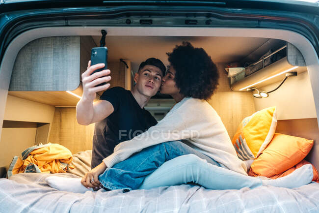 Young multiracial couple in love taking selfie on smartphone while hugging and kissing happily inside camper van during romantic journey together — Stock Photo
