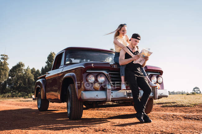 Cheerful couple of travelers orientating with paper map near red vintage pickup car parked on sandy road in summer — Stock Photo