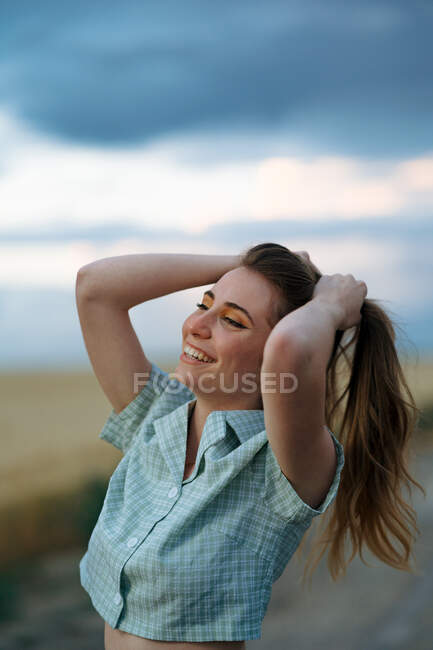 Счастливая стильная молодая женщина, прикасающаяся к длинным волосам, смотрит на дорогу под облачным небом в сумерках — стоковое фото