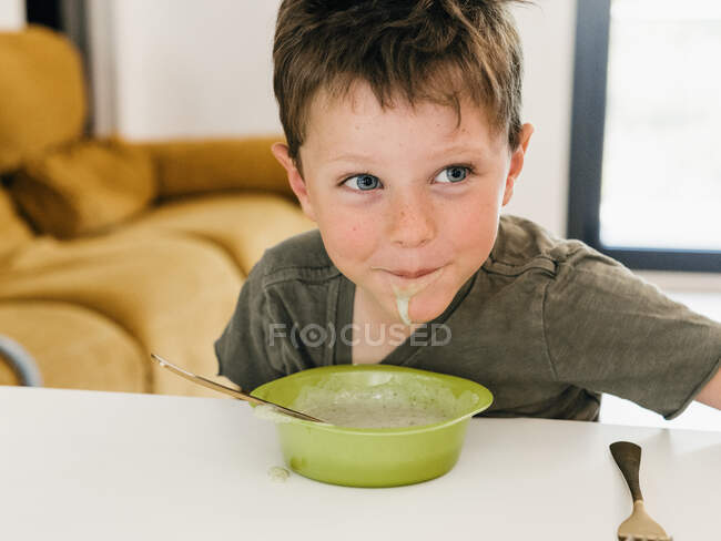 Ragazzo con la bocca sporca seduto a tavola con ciotola di zuppa di crema durante il pranzo e guardando altrove — Foto stock