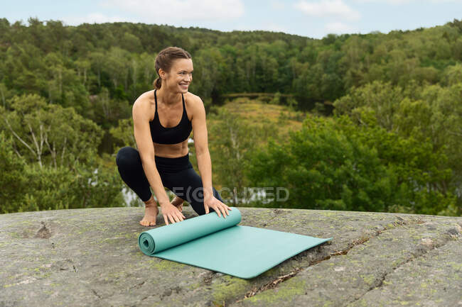 Mujer descalza de cuerpo completo en ropa deportiva negra está desenrollando la estera en la roca al inicio de la sesión de yoga cerca del pantano en la naturaleza - foto de stock