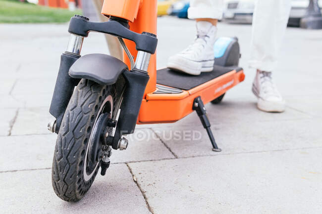 Cultivo persona anónima de pie cerca de scooter eléctrico estacionado en la calle en la ciudad durante el día - foto de stock