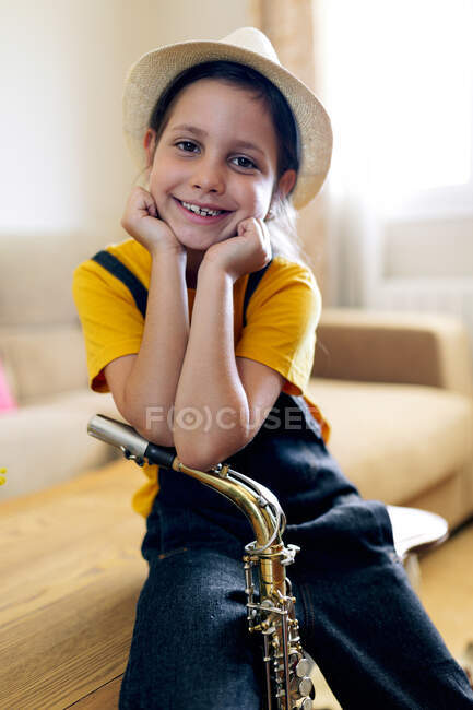Весела дитина в капелюсі з саксофоном дивиться на камеру, сидячи на столі в кімнаті — стокове фото