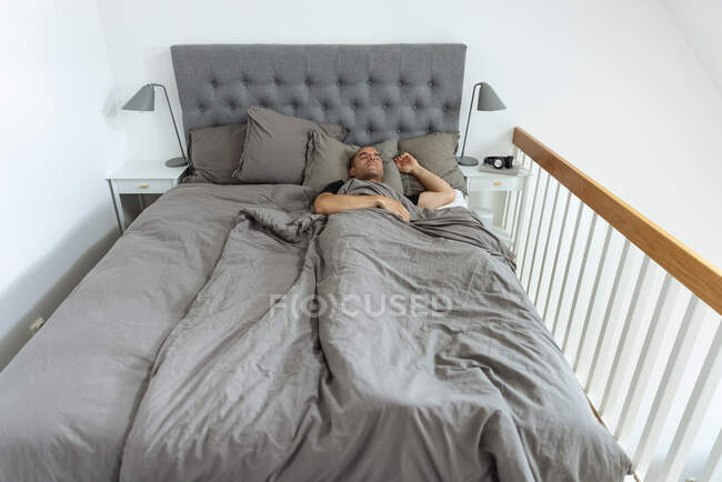 Сверху мирный мужчина лежит в мягкой постели под одеялом и спит утром в спальне — стоковое фото