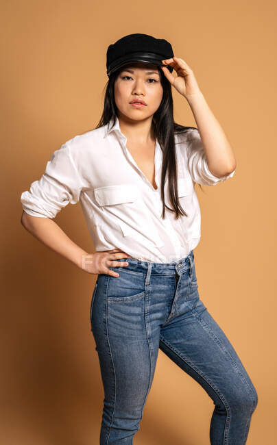 Front view of trendy Asian female model in white shirt and jeans touching cap on beige background and looking at camera — Stock Photo
