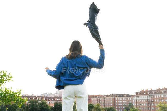 Обратный вид анонимной женщины в модной одежде, размахивающей шарфом на фоне неба в городе в ветреный день — стоковое фото