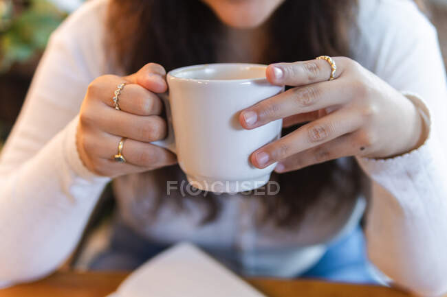 Cropped unrecognizable female enjoying delicious aromatic coffee from ceramic cup while resting in cozy cafe with green plants — Stock Photo