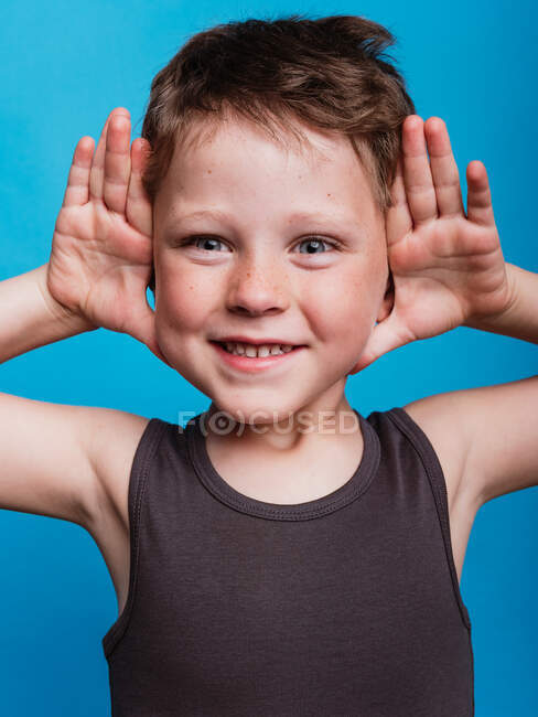 Joyful menino pré-adolescente com palmas abertas perto do rosto expressando felicidade no estúdio em fundo azul brilhante — Fotografia de Stock